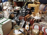 A collection of various vintage objects including cameras, bronze sculptures, picture frames, a stack of books, ornate jars, and busts are arranged on a table. The items are diverse, ranging from decorative to utilitarian, and create a sense of an antique market or collection.