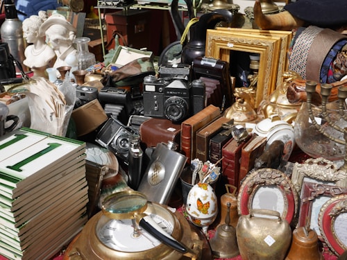 A collection of various vintage objects including cameras, bronze sculptures, picture frames, a stack of books, ornate jars, and busts are arranged on a table. The items are diverse, ranging from decorative to utilitarian, and create a sense of an antique market or collection.