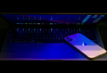 A smartphone with a glowing logo rests on the keyboard of a laptop. The scene is dimly lit, with a focus on blue and purple hues reflecting from the devices.