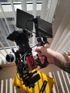 A professional video camera setup is being held by a person with a tattoo on their arm. The camera has various components including a lens, monitor, and cables. It is pointed towards a window with vertical blinds partially closed, allowing sunlight to filter through. The floor beneath displays the shadows of the blinds, and a yellow equipment case is visible.