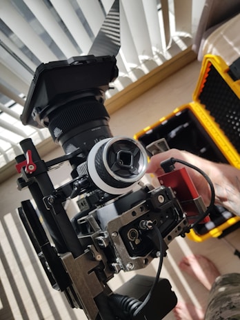 A professional video camera setup is placed on a flat surface near a window with partially open blinds. The camera is mounted on a stabilizing rig, and there is a monitor attached to it. A hand appears to be adjusting components of the camera setup. In the background, a yellow hard case is visible, likely used for transporting the equipment.