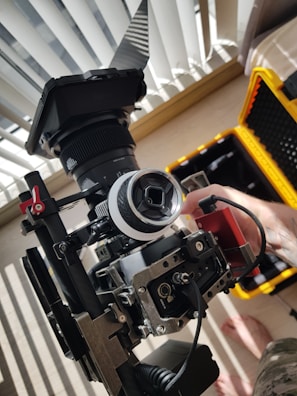 A professional video camera setup is placed on a flat surface near a window with partially open blinds. The camera is mounted on a stabilizing rig, and there is a monitor attached to it. A hand appears to be adjusting components of the camera setup. In the background, a yellow hard case is visible, likely used for transporting the equipment.