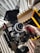 A professional video camera setup is placed on a flat surface near a window with partially open blinds. The camera is mounted on a stabilizing rig, and there is a monitor attached to it. A hand appears to be adjusting components of the camera setup. In the background, a yellow hard case is visible, likely used for transporting the equipment.