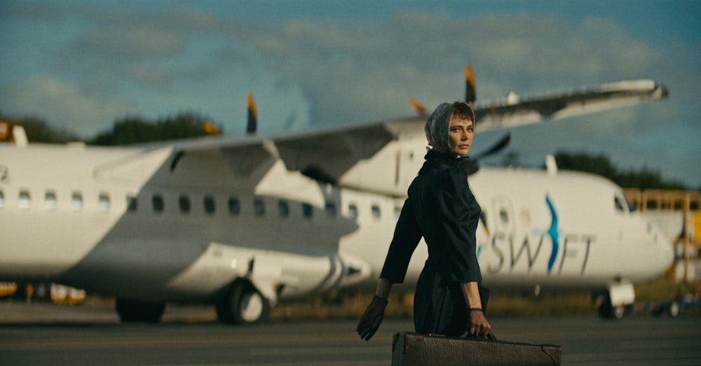 a woman in a black dress carrying a suitcase in front of an airplane