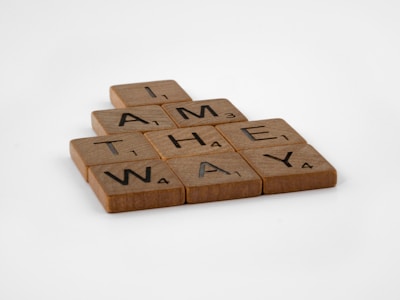 Wooden Scrabble tiles arranged to form the phrase 'I AM THE WAY'. Each tile displays a letter with an associated point value on a light, wooden surface.