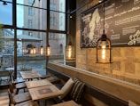 A cozy cafe interior featuring wooden tables, cushioned brown benches, and a large window showing an urban street view. The cafe is decorated with industrial-style hanging pendant lights and a large map wall art. The ambiance is warm and inviting.