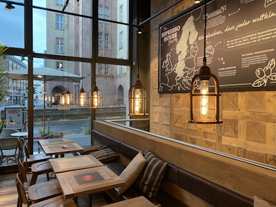A cozy cafe interior featuring wooden tables, cushioned brown benches, and a large window showing an urban street view. The cafe is decorated with industrial-style hanging pendant lights and a large map wall art. The ambiance is warm and inviting.