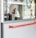 Two individuals wearing red caps and reflective vests, likely security or service personnel, stand near a large glass door. One person is holding a small object, possibly a radio or scanner. Reflections in the glass show parts of office interiors and posters. There are additional people sitting inside, visible through the glass.