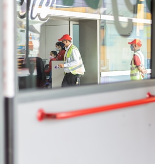 Two individuals wearing red caps and reflective vests, likely security or service personnel, stand near a large glass door. One person is holding a small object, possibly a radio or scanner. Reflections in the glass show parts of office interiors and posters. There are additional people sitting inside, visible through the glass.
