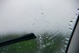 A close-up view of a windshield with raindrops being wiped by a windshield wiper. The wiper blade is in the middle of a swipe, clearing a path through the droplets.