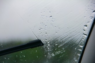 A close-up view of a windshield with raindrops being wiped by a windshield wiper. The wiper blade is in the middle of a swipe, clearing a path through the droplets.