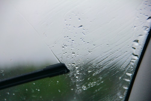 A close-up view of a windshield with raindrops being wiped by a windshield wiper. The wiper blade is in the middle of a swipe, clearing a path through the droplets.