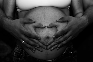 A close-up of hands gently resting on a pregnant belly, with two pairs of hands forming a heart shape. The image is in black and white, highlighting the textures and contrast.