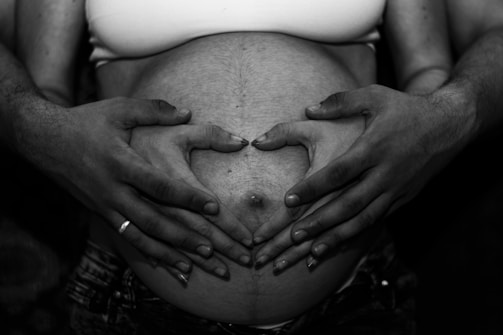 A close-up of hands gently resting on a pregnant belly, with two pairs of hands forming a heart shape. The image is in black and white, highlighting the textures and contrast.