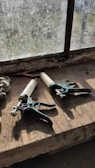 Two caulking guns with white cartridges and black handles are placed on a dusty windowsill. The window is old and dirty, partially obstructing the view outside. A few small tools and a rag are also visible on the windowsill.