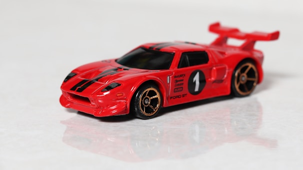 A red toy car model with black racing stripes and a large rear spoiler. It features gold-colored wheels and a racing number one on the side. The car resembles a sports car design and is positioned on a reflective surface.