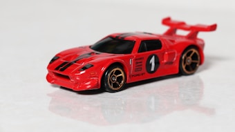 A red toy car model with black racing stripes and a large rear spoiler. It features gold-colored wheels and a racing number one on the side. The car resembles a sports car design and is positioned on a reflective surface.