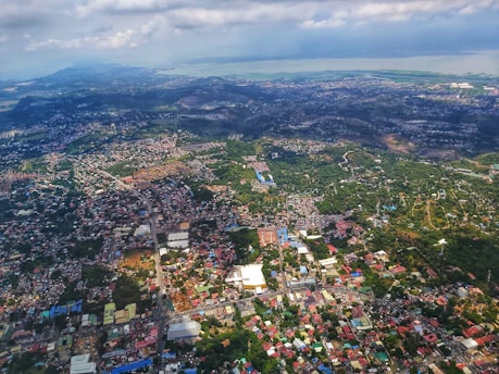 An aerial view of a densely built cityscape with a mix of residential and commercial areas. The city is surrounded by green hills and stretches towards the coast in the background. Buildings are clustered together, and the landscape features a combination of urban development and natural green space.