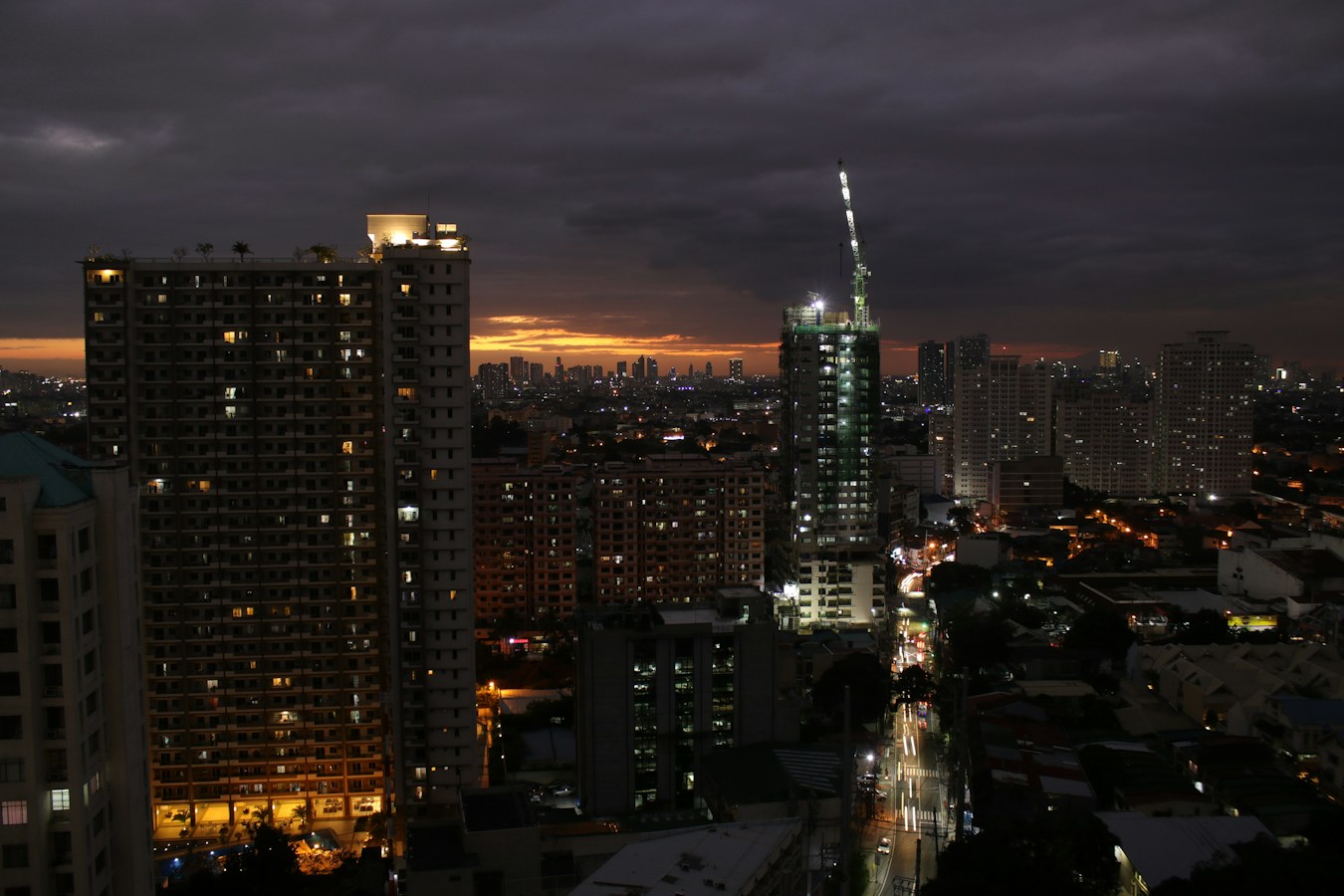 A Manila high-rise cityscape at dusk with lit residential towers, a crane-topped tower under construction, and a lit street cutting through the city