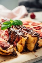 A close-up of a dessert featuring layers of pastry topped with rich chocolate sauce and garnished with a fresh basil leaf. The pastry appears to be filled with a red fruit, possibly strawberry or cherry. The image creates a warm and inviting atmosphere with a soft focus on the background.