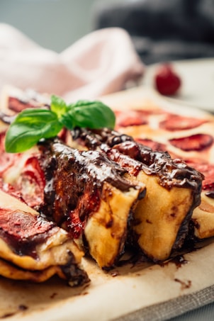 A close-up of a dessert featuring layers of pastry topped with rich chocolate sauce and garnished with a fresh basil leaf. The pastry appears to be filled with a red fruit, possibly strawberry or cherry. The image creates a warm and inviting atmosphere with a soft focus on the background.
