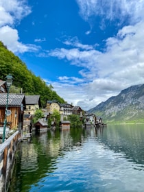 A picturesque lakeside village with charming houses nestled along the water's edge. The vibrant greenery of the surrounding trees contrasts with the clear blue sky above, while the calm water reflects the serene landscape. Majestic mountains rise in the background, adding to the scenic beauty of the location.