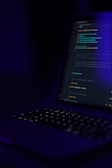 A dimly lit laptop keyboard and screen displaying lines of code against a dark background. The focus is on the coding text, which is illuminated by the screen's glow, highlighting elements such as functions and syntax in different colors.