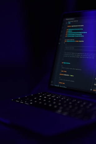 A dimly lit laptop keyboard and screen displaying lines of code against a dark background. The focus is on the coding text, which is illuminated by the screen's glow, highlighting elements such as functions and syntax in different colors.