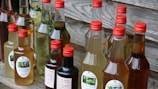 A collection of various bottles filled with different colored liquids, each with a red cap, displayed on a wooden surface. Some bottles have labels with text and images, suggesting homemade or artisanal products.