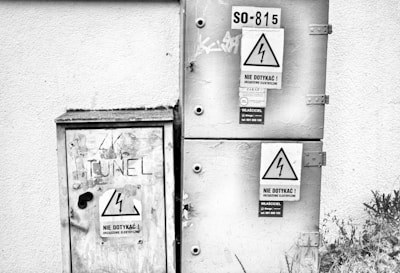 A black and white image featuring two electrical boxes mounted on a wall. The boxes have warning signs with the lightning bolt symbol, indicating high voltage, and text in Polish cautioning not to touch. Graffiti and minor scratches are visible on the surfaces. The environment appears industrial and slightly weathered.