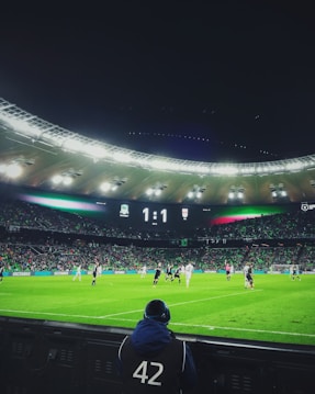 A soccer match taking place in a large stadium filled with spectators. The scoreboard shows a score of 1:1, and players dressed in dark and light jerseys are actively engaged on the well-lit, green field. A person wearing a jacket with the number 42 can be seen in the foreground, facing the field.