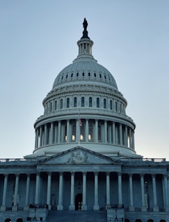 A grand neoclassical building with a large dome and columns, featuring an American flag in front. It appears to be a government structure with detailed architectural features.