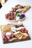 Three boards filled with an assortment of foods are placed on a white surface. The boards are laden with fresh strawberries, grapes, blueberries, assorted nuts, crackers, meats, cheese, and various other snacks. Vibrant flowers also adorn the boards, adding a touch of color and decorative flair.