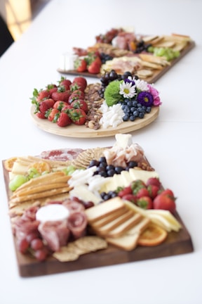 Three boards filled with an assortment of foods are placed on a white surface. The boards are laden with fresh strawberries, grapes, blueberries, assorted nuts, crackers, meats, cheese, and various other snacks. Vibrant flowers also adorn the boards, adding a touch of color and decorative flair.