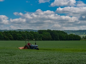 A vast green field stretches out with a lone blue tractor steadily making its way across the land. The backdrop features lush green trees with rolling hills visible in the distance. Above, fluffy clouds are scattered across a vibrant blue sky.