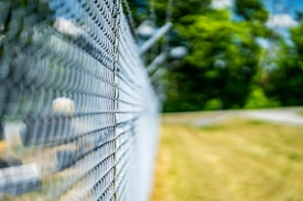 A close-up view of a chain-link fence with a shallow depth of field, creating a blurred background. The fence stretches diagonally across the image from the foreground to the distance. Behind it, vibrant green trees and a grassy field are partially visible under a bright blue sky.