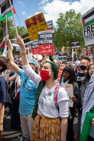 A group of people participate in a protest or demonstration, holding signs advocating for Palestine. Many in the crowd wear face masks, and a prominent sign reads 'Freedom for Palestine.' The atmosphere is energetic and unified with participants raising their fists in solidarity.