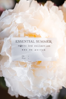 A white, fluffy flower with a perfume label attached to it. The label reads 'ESSENTIAL SUMMER' and includes details about a perfume collection. The background is blurred, focusing attention on the flower and the label.