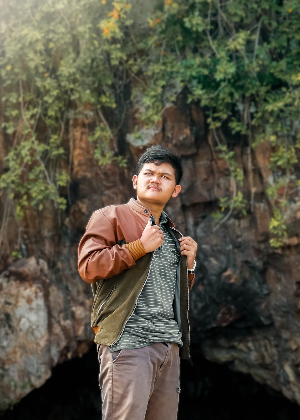 In the image, a young man is standing against a backdrop of a large rock formation and lush green foliage. The man is dressed casually for the occasion, wearing a brown jacket over a gray shirt paired with khaki pants. He holds his hands in his pockets while looking up at something above him.
