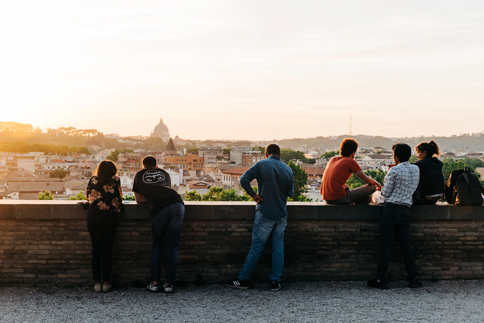 Travelers gathered along a garden wall in Rome at golden hour, looking out over the rooftops toward St. Peter's dome.