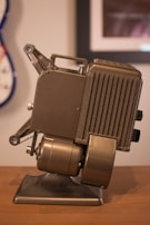 A vintage metal film projector with a metallic finish rests on a wooden surface. The device features a classic design with dials, a vented side panel, and mechanical parts. The background is slightly blurred, showing part of a wall with framed photos and a guitar partially visible.
