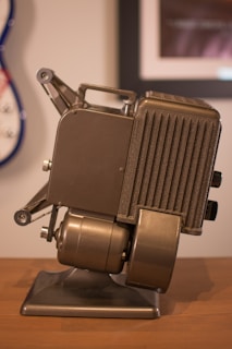 A vintage metal film projector with a metallic finish rests on a wooden surface. The device features a classic design with dials, a vented side panel, and mechanical parts. The background is slightly blurred, showing part of a wall with framed photos and a guitar partially visible.
