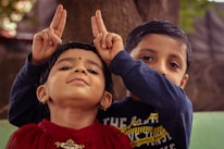 Two young children are engaging in playful antics. The child in the foreground, wearing a red top, has a confident expression and is making a playful gesture resembling rabbit ears behind their head with both hands. Behind them, another child can be seen with a curious expression, wearing a dark-colored shirt. They are against a natural outdoor background.