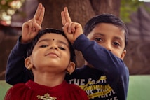 Two young children are engaging in playful antics. The child in the foreground, wearing a red top, has a confident expression and is making a playful gesture resembling rabbit ears behind their head with both hands. Behind them, another child can be seen with a curious expression, wearing a dark-colored shirt. They are against a natural outdoor background.