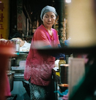 A woman wearing a pink shirt and a gray headscarf is working at a food stall. She appears focused and is possibly preparing food. The surroundings are a bit cluttered with various containers and cooking items. The setting seems to be a bustling market or street food area with dim lighting.