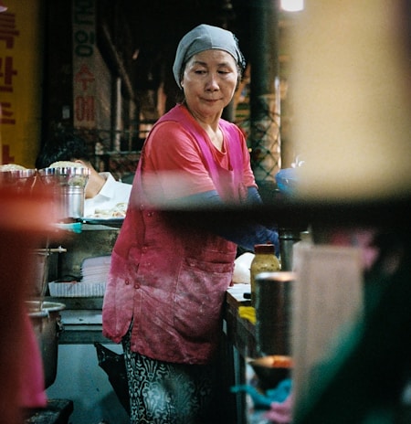 A woman wearing a pink shirt and a gray headscarf is working at a food stall. She appears focused and is possibly preparing food. The surroundings are a bit cluttered with various containers and cooking items. The setting seems to be a bustling market or street food area with dim lighting.