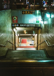 An underground subway entrance with stairs leading down into a station. The area is illuminated with fluorescent lights and there are signs in Korean and English. The walls are lined with tiles, and there are handrails on either side of the stairs. The scene appears to be in an urban environment with some visible street elements at the top.