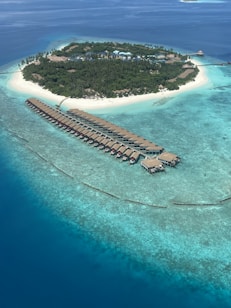 An aerial view of a tropical island surrounded by a pristine turquoise ocean. The island is densely covered with green vegetation, and there are multiple buildings with thatched roofs. Extending from the island into the water is a long series of overwater bungalows arranged in a curved pattern. The water transitions from shallow light blue near the island to deeper dark blue further out.