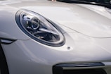A close-up of a sleek, modern car's front headlight and part of the hood. The headlight design is intricate and reflects light, with a clear lens covering the bulb and reflective elements. The car's surface is glossy, suggesting a polished, pristine condition.