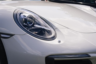 A close-up of a sleek, modern car's front headlight and part of the hood. The headlight design is intricate and reflects light, with a clear lens covering the bulb and reflective elements. The car's surface is glossy, suggesting a polished, pristine condition.